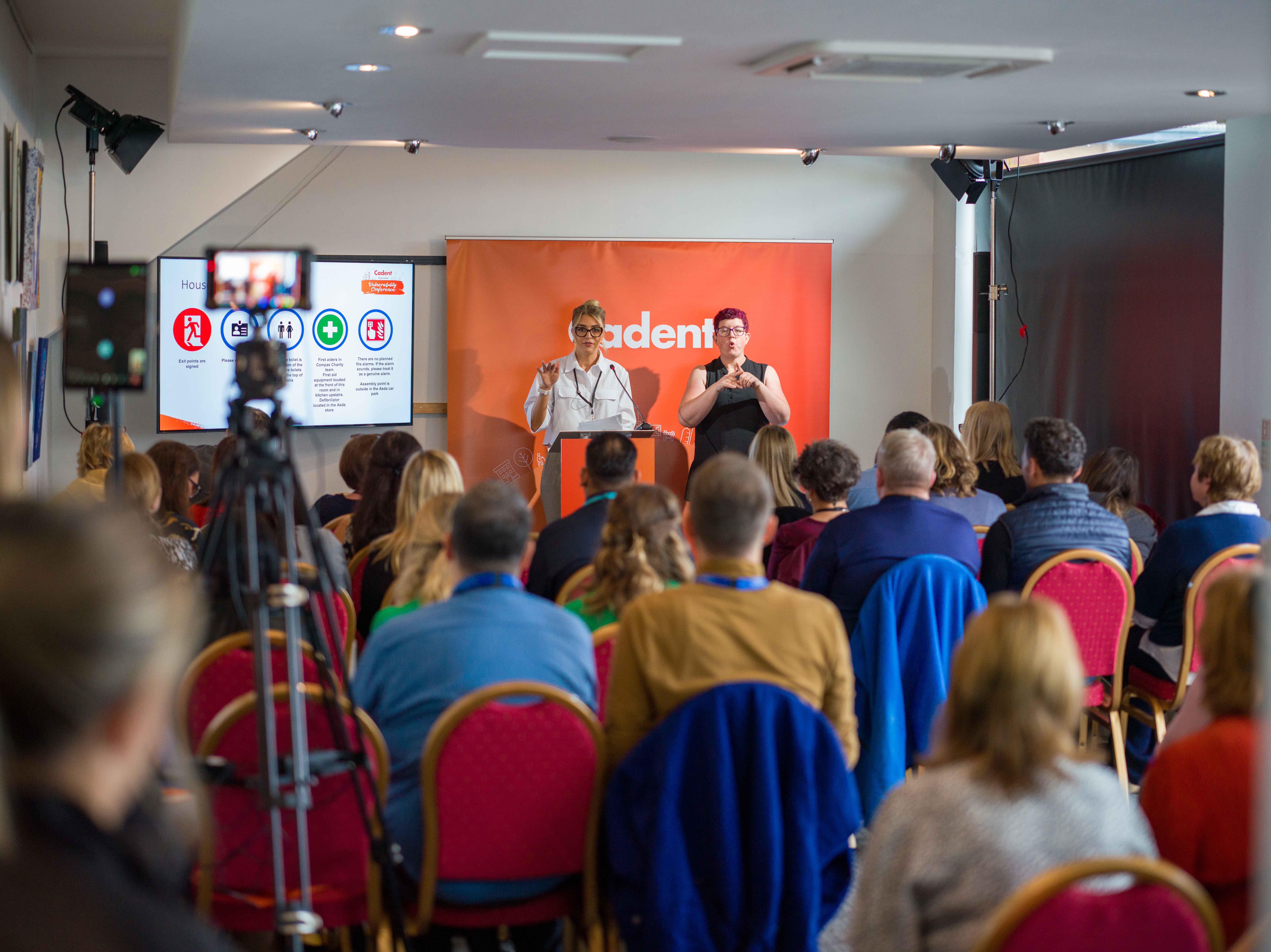 Presenters addressing a diverse audience at a community support conference, with sign language interpretation on stage.