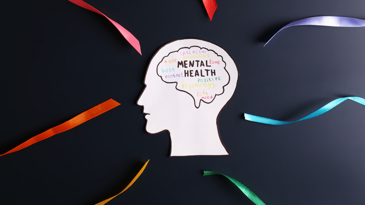 Illustration of a human head silhouette with colorful mental health and wellness words inside the brain area, surrounded by multicolored ribbons on a dark background.