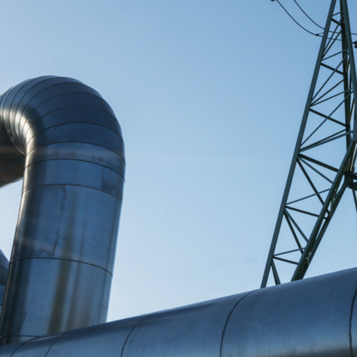 Large metallic industrial pipes with a bend rise against a clear blue sky, accompanied by a green metal lattice structure. The scene feels industrial and expansive.