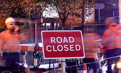 Workers in orange vests stand near a 'Road Closed' sign at night. Barriers and blurred motion convey a bustling, active work scene.