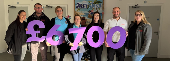 A group photo related to a charity activity, showing people holding large £6700 signs indoors, with a visible Jolly Josh charity poster displayed in the background