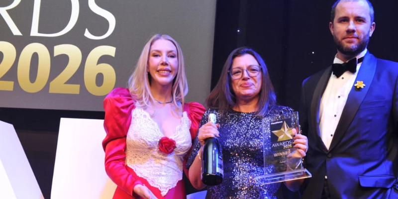 A group of individuals dressed in formal attire stand onstage at an awards event, holding a trophy and a champagne bottle against a backdrop displaying Fleet News Awards 2026 in bold lettering.