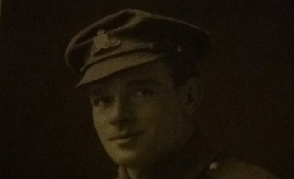 A sepia portrait of a soldier wearing a peaked military cap with an emblem. The soldier presents a gentle smile, conveying a solemn and respectful tone.
