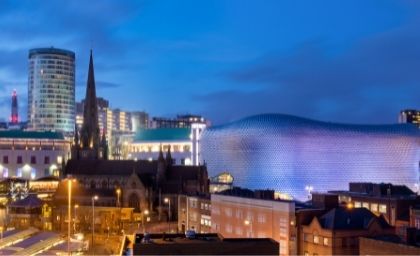 Cityscape at dusk featuring modern and historic architecture. A futuristic building with a textured facade is illuminated beside a church and lit-up skyscrapers.