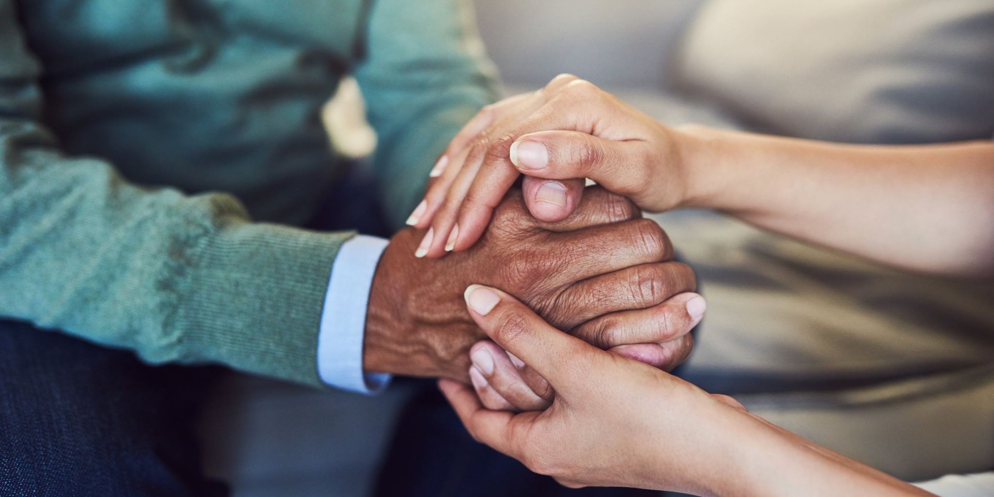 Close-up of two people gently holding an older person's hands, conveying care, comfort, compassion, and emotional support.