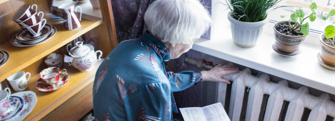 An elderly woman in a blue robe inspects a radiator while holding papers. Houseplants sit on the windowsill, and a cabinet displays teacups nearby.