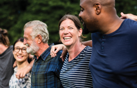 A diverse group of five people stands arm in arm outdoors, sharing smiles and enjoying each other's company in a green park setting.