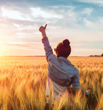A woman wearing a denim jacket stands in a golden wheat field, joyfully raising her hand toward a colourful sunset sky.